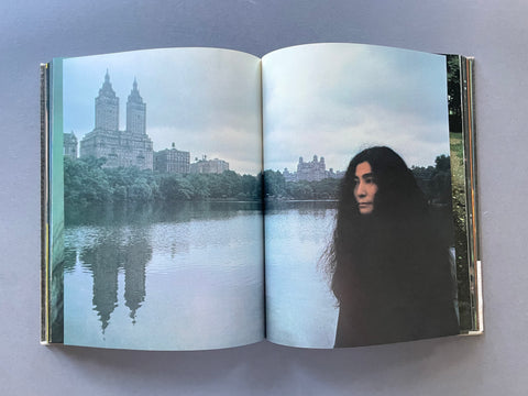 An open hardcover book laid flat on a gray surface, showing a printed photograph spread across both pages. The photo depicts Yoko Ono with long dark hair standing by a calm lake with a large historic building and trees in the background.