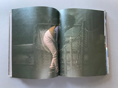 An open book or magazine lying flat on a light gray surface, showing a large photographic print on the spread with a dark, moody scene of a person in light-colored clothing near a fence.