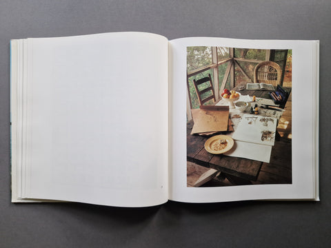An open hardcover book, showing a mostly blank white left page and a printed interior photograph on the right page depicting a rustic wooden table with dishes and papers in a sunlit room.
