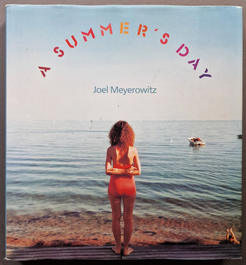 The photobook cover of Joel Meyerowitz's A Summer's Day with a photo of a red head woman in a red swimsuit with her back to the camera on a shore.