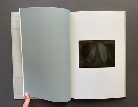 An open book or photo album on a dark gray background, showing a large pale blue-gray page turned open and a small dark square photograph printed on the right page. A hand is visible holding the lower left edge of the page.