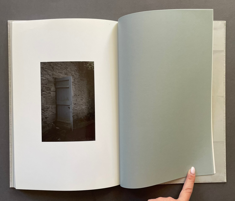 An open art book on a dark gray surface, showing a white page with a small dark photograph of a door and a pale sage-green page being turned by a hand.