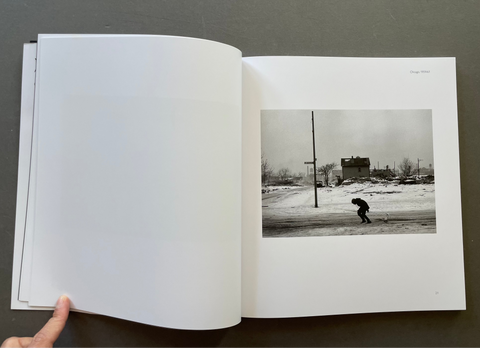 An open book or photo journal lying on a dark gray surface, with a hand holding the left page. The right page shows a black-and-white landscape photograph of a person walking through a snowy outdoor scene.