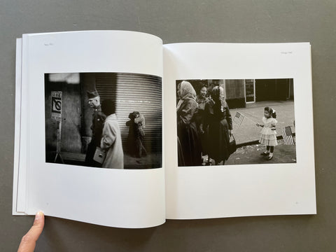 An open book or photo journal laid flat on a dark gray surface, showing two printed black-and-white photographs spread across the pages. A human hand is visible holding the left edge of the book.