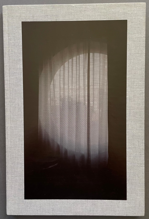A square framed photo book featuring a photograph of a soft light glowing through sheer curtains in an arched window, set within a textured off-white linen-like border, displayed against a dark gray wall.