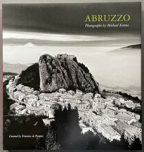 A hardcover art book titled 'ABRUZZO: Photographies by Michael Kenna' lying flat on a textured gray surface. The cover features a black and white landscape photograph of a town perched on a rocky hill, with the title printed in yellow at the top right.