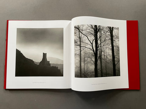 An open hardcover art book lying flat on a gray surface, displaying two black and white landscape photographs side by side. The left page shows a dark, moody scene of a castle ruin under a cloudy sky, and the right page shows bare trees in mist.