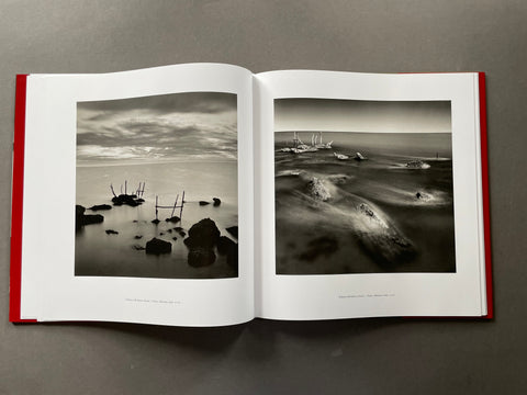 An open hardcover art book with red side borders, displaying two black-and-white landscape photographs on white pages against a dark gray background.
