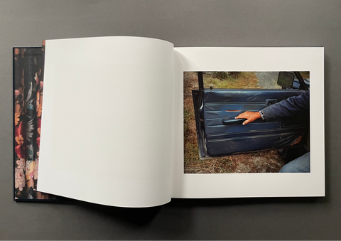 An open hardcover book or photo journal lies on a dark gray surface. The left page is blank white, while the right page shows a printed photograph of a hand reaching for a car door handle.