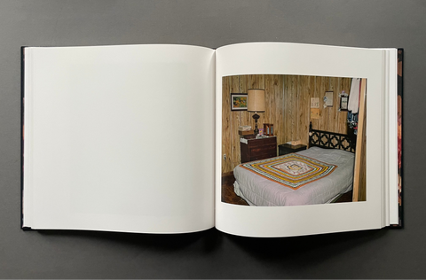 An open hardcover book or photo album laid flat on a dark gray background, showing a printed interior bedroom photograph on the right page with blank white pages on the left.