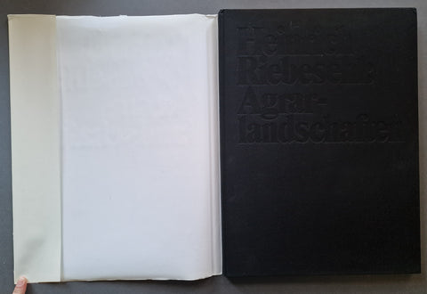 An open book showing a large black embossed cover with the title 'Heinrich Riebesehl Agrarlandschaften' and a white inner page or folder beside it, photographed against a dark gray surface.