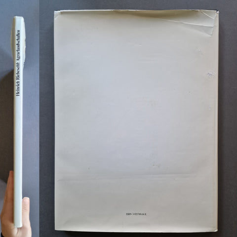 A hand holds a slim white book or art print next to a larger off-white square sheet or box on a dark gray background. The spine shows the text 'Heinrich Riebesehl Agrarlandschaften' and the bottom of the square piece shows 'RIN.