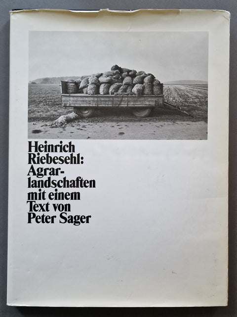 A book cover for 'Heinrich Riebeschl: Agrar-landschaften mit einem Text von Peter Sager' featuring a black and white photograph of a cart loaded with sacks on a dirt road, set against an off-white background.