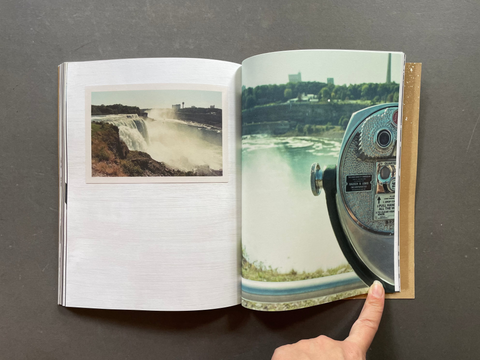 An open photo book or magazine lies on a dark gray surface, showing printed photographs of a waterfall and a coin-operated viewing bin. A person's finger is pointing at the right-hand page.