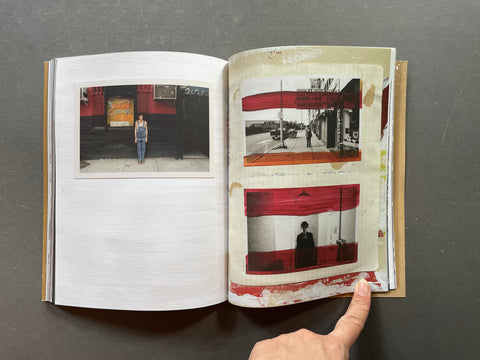 An open art book or photo journal lying on a dark gray surface, showing printed photographs and collage-style pages with prominent red and beige tones. A person's hand is visible holding the lower right edge of the book.
