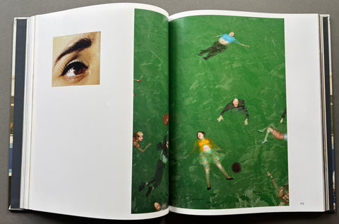 An open hardcover book spread on a dark gray surface, featuring a large green photographic print with multiple small human figures and a smaller inset photo of an eye on the left page.