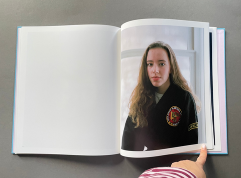 An open hardcover book lying on a gray surface, showing a printed photograph of a young woman in a dark jacket on the right page.