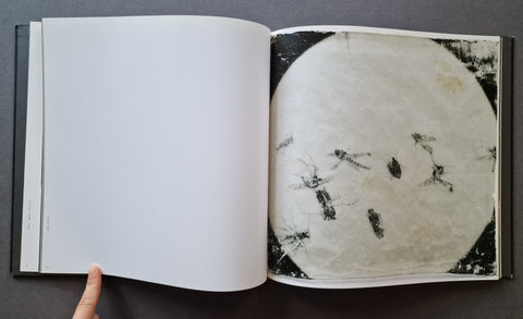 An open art book on a dark gray surface, showing a blank white page on the left and a printed artwork on the right with a large circular off-white form and black insect-like marks.