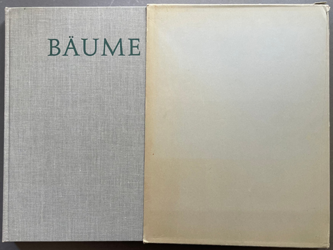 A vintage hardcover book or album titled 'BÄUME' on the left, featuring a textured gray linen cover with dark green lettering, next to a blank cream-colored dust jacket or inner cover on a dark background.