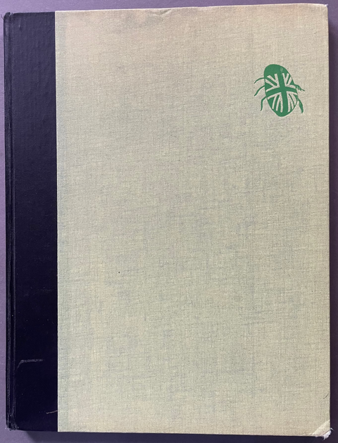 A vintage hardcover book or notebook with a textured off-white linen cover, a black spine band on the left, and a small green beetle emblem in the upper right corner, photographed flat against a dark background.