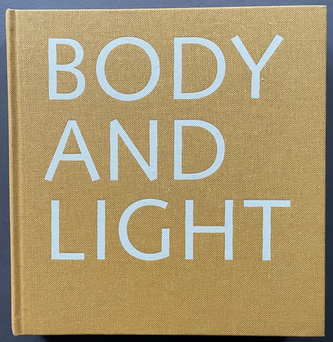A square hardcover book with a textured tan linen cover and large light blue sans-serif text reading 'BODY AND LIGHT' on the front, photographed against a dark background.