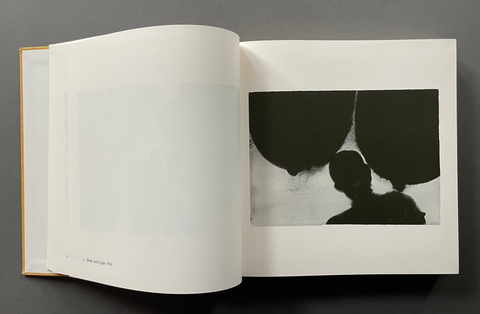 An open art book or magazine laid flat on a dark gray surface, showing a mostly blank left page and a right page with an abstract black-and-white photographic print of a silhouetted figure against dark shapes.