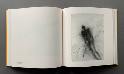 An open art book displayed flat against a dark gray background, showing a white page on the left with faint markings and a monochrome abstract artwork on the right featuring a dark charcoal figure on a light gray background.