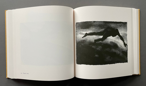 An open book laid flat on a dark surface, showing a black-and-white art print on the right page with a silhouetted human figure against a textured dark background, and mostly blank off-white pages on the left.