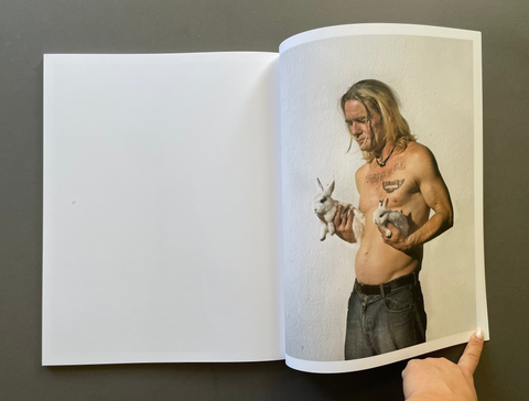 An open book lying on a dark gray surface, showing a printed photograph on the right page of a shirtless man holding two rabbits. A human hand is visible at the bottom right holding the page open.