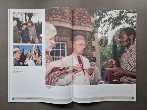 An open magazine spread photographed from above on a dark gray surface. The pages contain multiple photographs: a large main photo of people outdoors near a stone building, two smaller inset photos on the left, and small caption text at the bottom.