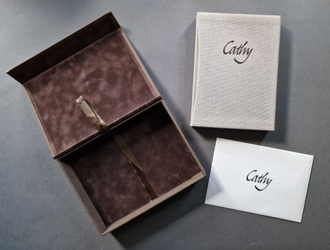 A limited edition box set featuring an open dark brown velvet-lined presentation box with a ribbon, a matching textured cream book titled Cathy in a slipcase, and a white card, all personalized with the name 'Cathy' in black script, arranged on a dark gray surface.