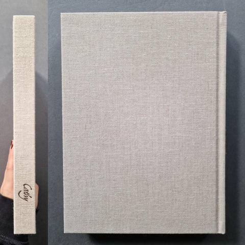 A hardcover book with a light beige linen-textured cover and matching spine, shown from the front and side on a dark gray background. The spine features the brand name 'Cathy' in black script.