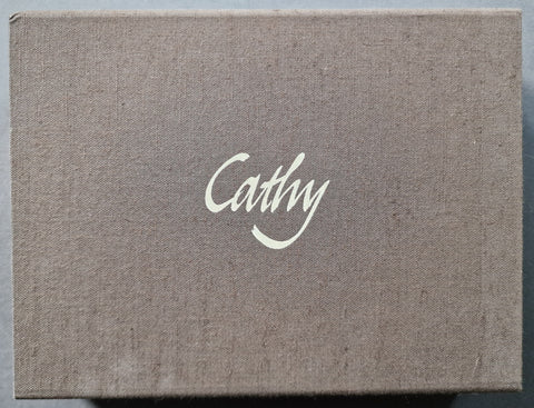 A square fabric-covered box with a textured taupe-brown linen-like surface and the name 'Cathy' printed in white script at the center.