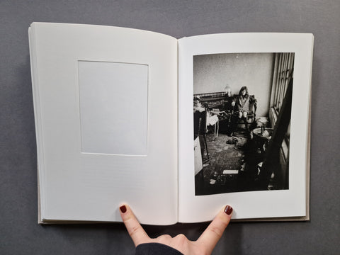 An open hardcover book: The left page has a blank white mat and inset frame, while the right page shows a black-and-white photograph of a cluttered interior scene and a girl.
