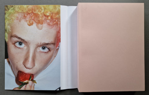 An open book, showing a close-up portrait of a person with curly yellow-orange hair biting a strawberry on the left page.