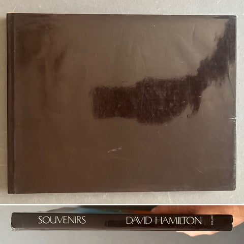 A hardcover book titled 'Souvenirs: David Hamilton' lying on a light gray surface, with the large front cover shown above and the spine visible below where a hand is holding it.