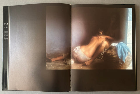 An open book or magazine spread showing a dark-toned interior spread with a large photograph on the right page of a crouching nude woman beside a basket, with a blue cloth draped over it.