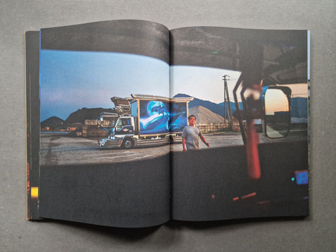 An open book photographed from above on a gray surface, showing a printed photograph of a truck with a large blue circular graphic on the side and a person standing nearby.