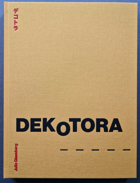 A hardcover book titled 'DEKOTORA' by Julie Glassberg, featuring a textured yellow linen cover with bold black typography and red vertical text, photographed flat against a blue background.