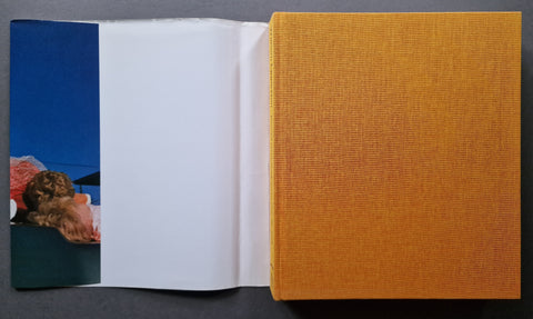 An open book with a textured mustard-yellow cover on the right, white inner pages in the center, and a blue-toned printed page on the left, photographed against a dark gray background.