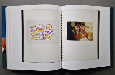 An open hardcover book, showing two interior pages with black binding strips. The left page contains a small abstract artwork in purple, yellow, and orange lines on a white sheet, and the right page contains a small color photograph of a candlelit table scene.