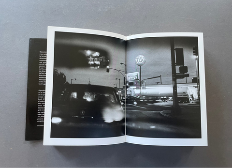 An open black-and-white art book or magazine lying on a gray surface, showing a street scene photograph across both pages with a dark cover visible on the left.