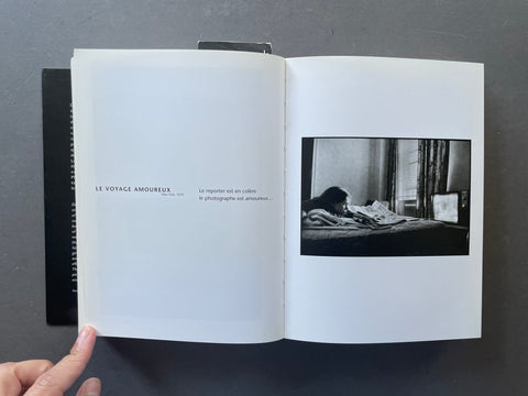 An open book lying on a dark gray surface, showing a black-and-white interior photograph printed on one page and French text on the other. A person's fingers are visible holding the left page open.