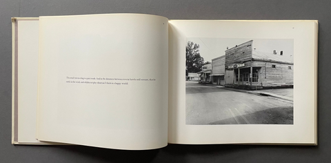 An open book or art print laid flat on a dark gray surface, showing an off-white page with small printed text on the left and a black-and-white photograph of a street scene with buildings on the right.