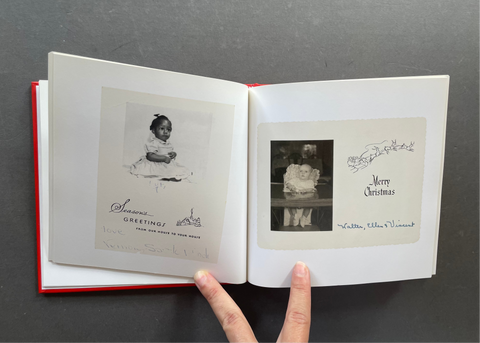 An open vintage photo album or book with two printed pages, held open by a person's fingers against a dark gray background. The pages show black-and-white photographs with handwritten holiday greetings and decorative text.