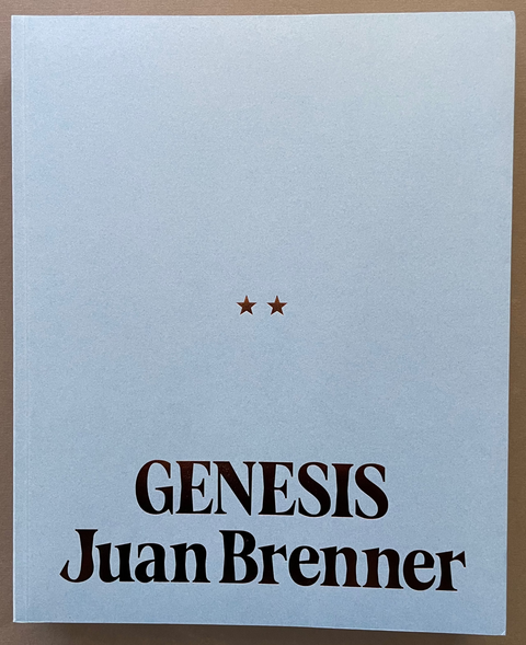 A book cover for 'Genesis' by Juan Brenner, featuring a minimalist light blue-gray background with two small stars and bold black text, photographed flat against a brown surface.