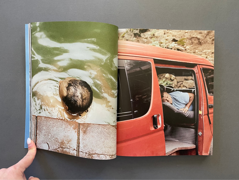 An open printed magazine or book lying on a dark gray surface, showing two photographic pages side by side. The left page has a green-tinted photo of a person in shallow water, and the right page shows a red-orange van with a person reclining inside.