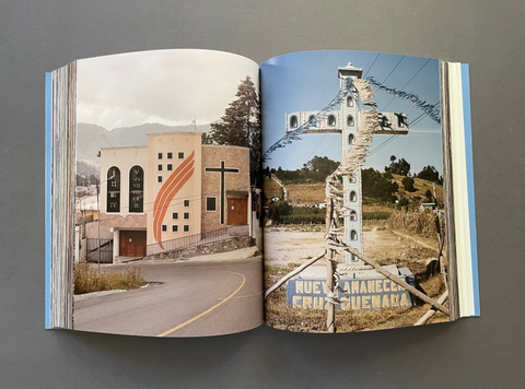 An open book or photo journal laid flat on a gray surface, showing two printed photographs spread open. The left page depicts a beige church building with a cross and mountain background, and the right page shows a tall utility pole structure in an outdoor rural setting with the text 'RUE ESTRELLA FRUTILLA GUENAGA'.
