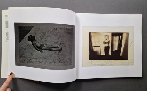 An open art book. The left page shows a black-and-white photograph of a person lying on sand with handwritten text, and the right page shows a sepia-toned photograph of a person standing in a doorway with handwritten text below.
