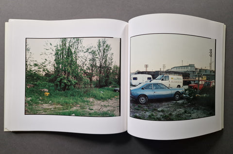 An open hardcover book, showing two printed photographs on the spread. The left page contains a landscape photo with trees and grass, and the right page shows a street scene with parked cars and buildings.